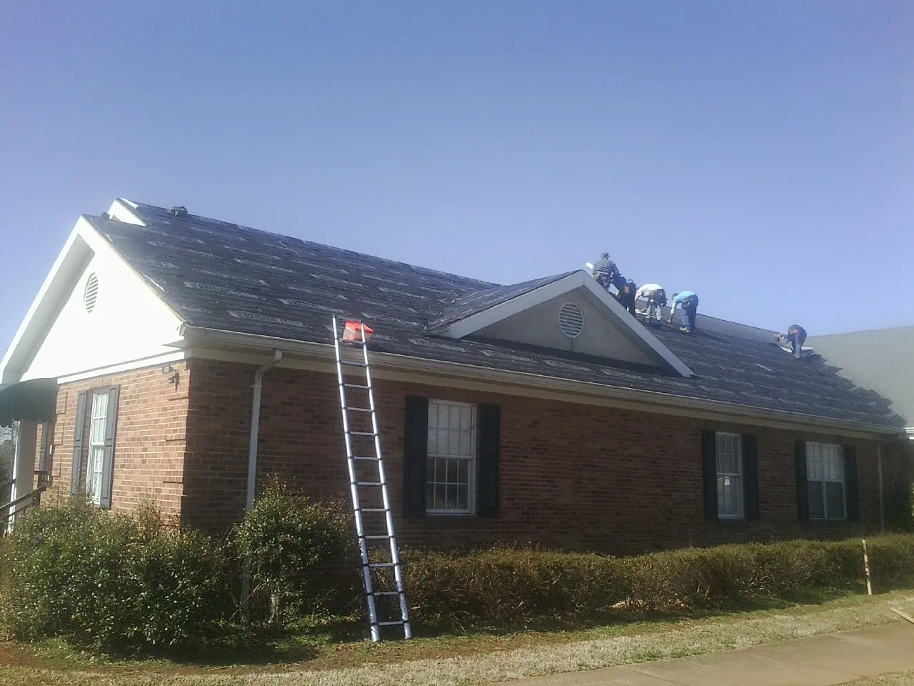 Crew tearing shingles off a brick ranch roof with a ladder set — Emergency Roof Repair work by our crew in Hollis